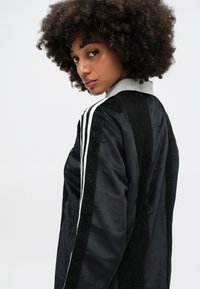 Jachetă din satin negru cu dungi albe pe umeri, panouri texturate negre și guler alb. Prezintă un design elegant și sportiv.