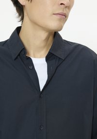 Chemise bleu marine à manches longues avec col boutonné, coutures apparentes et deux boutons au niveau du cou. Portée sur un t-shirt blanc.