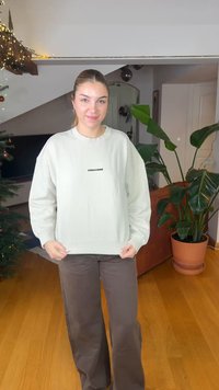 Felpa oversize verde chiaro con polsini e orlo a coste, caratterizzata da un logo nero sul davanti. Abbinata a pantaloni larghi marroni.