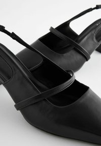 Tacones de piel negra tipo slingback con punta afilada, con una amplia correa cruzada y una textura suave. El tacón es de altura media.