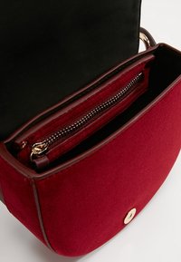 Červená látková crossbody taška se zlatým knoflíkem a zipovou kapsou; hladká textura s hnědými akcenty vystýlajícími interiér i exteriér.