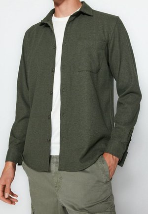 Homme portant une chemise boutonnée vert olive sur un t-shirt blanc avec un pantalon cargo olive assorti, main dans la poche, debout devant un fond blanc.