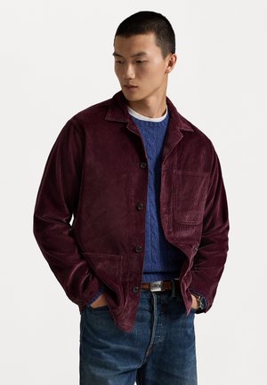 Veste en velours côtelé bordeaux avec une fermeture à bouton, une poche poitrine simple et un col cranté, portée par-dessus un pull en mailles torsadées bleu et un jean.