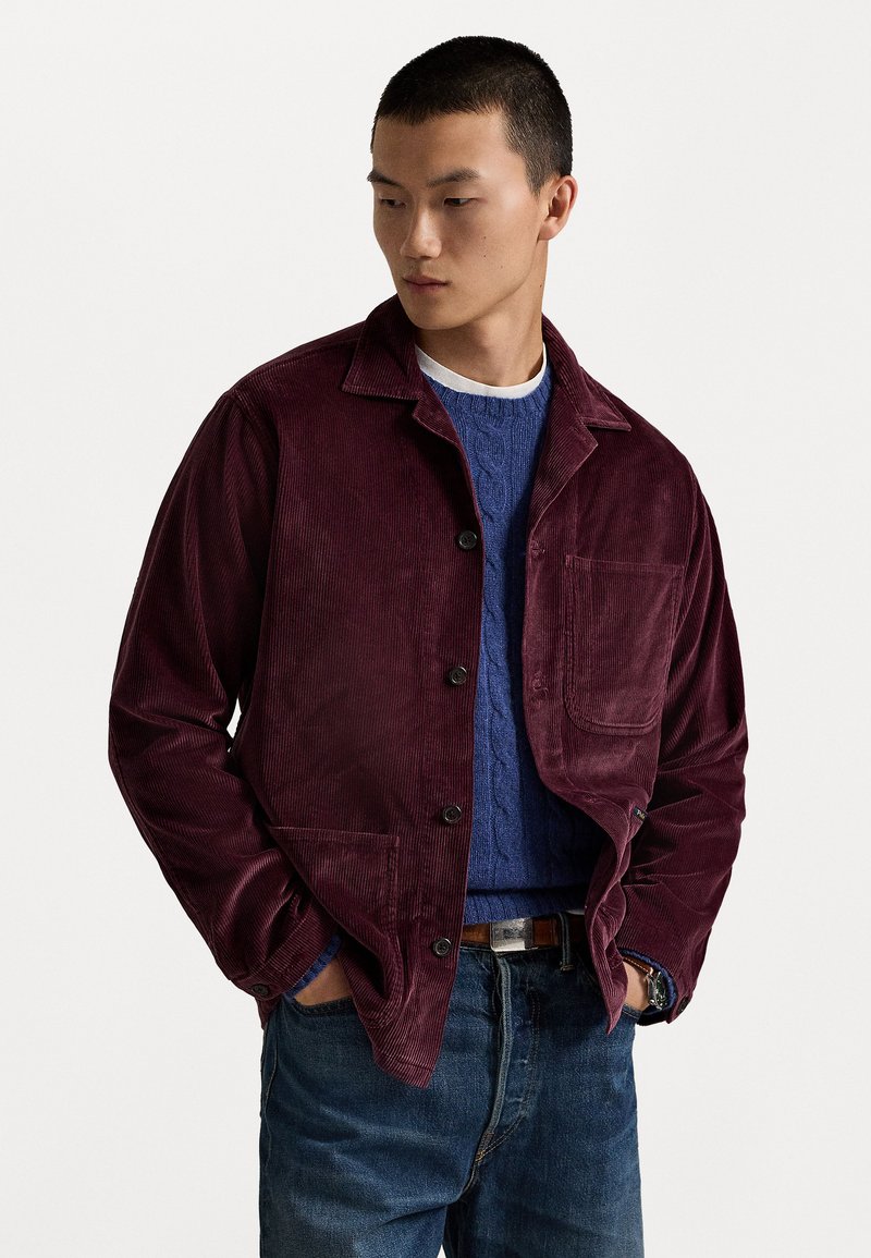 Burgundy corduroy jacket with a button closure, single chest pocket, and notch collar, layered over a blue cable-knit sweater and jeans.