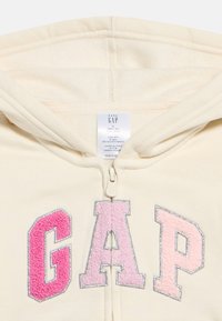 Béžová detská mikina s kapucňou a zipsom, na prednej strane s nápisom "GAP" v ružovej textúrovanej farbe a s kapucňou navrchu.
