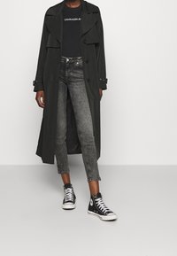 Abrigo negro tipo trench con cierre de botones y cuello ancho, combinado con jeans ajustados grises y zapatillas de caña alta negras. Tejido suave, estilo casual.