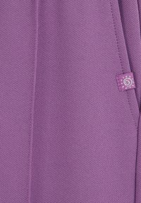 Tuvaļu violeta faktūra audums ar centrālo šuvi un nelielu piešūtu etiķeti, kurā attēlots sudraba spirāles saules dizains.