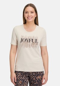 T-shirt beige à manches courtes avec un col rond, ornée de "JOYFUL" en écriture cursive noire et de décorations métalliques dorées. Associé à un pantalon à motifs.