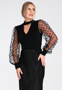Czarny top z dekoltem w serek, krótka, gładka góra i przezroczyste bufiaste rękawy w kropki. Połączony z dopasowaną czarną spódnicą. Gładka tekstura.