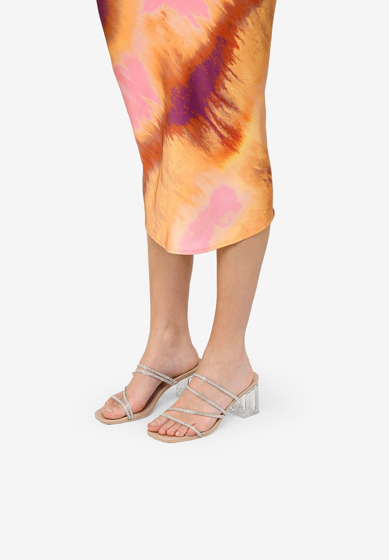 Sandalias con tiras plateadas y un tacón bloque transparente, que presentan una suela nude. Combinadas con un vestido colorido de patrón abstracto.