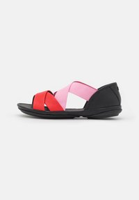 Svart sandal med röd och rosa elastisk korsrem, rund tå, flexibel sula och texturerade läderdetaljer.