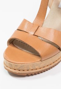 Gros plan d'une sandale en cuir beige avec une semelle épaisse et à motifs, et de larges brides croisées sur les orteils.