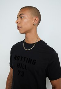 Svart T-skjorte med "NOTTING HILL 73" i fet, teksturert skrift. Personen har på seg en sølvkjede og hoop-øreringer. Huden er lysbrun.