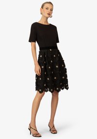 Robe noire avec un haut ajusté et des manches courtes, présentant une jupe florale texturée avec des accents beiges et un ourlet festonné.