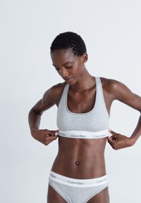 Light grey cotton sports bra with a wide white band featuring "Calvin Klein" logo; paired with matching underwear. Smooth texture, minimalist design.