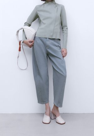 Ljusgrön figurnära jacka med hög krage, matchad med lösa ljusblå byxor och krämfärgade klackskor med blockklackar, hållandes en vit handväska.