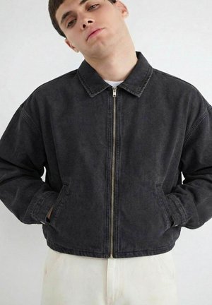 Mladý muž v čiernej vetrovke na zips s golierom, ruky v vreckách, stojí na jednoduchom svetlom pozadí.