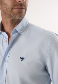Hellblaues Hemd mit Knöpfen, das eine weiche Textur, einen Kragen und ein kleines, navyblaues, gesticktes Vogel-Detail auf der linken Seite aufweist.