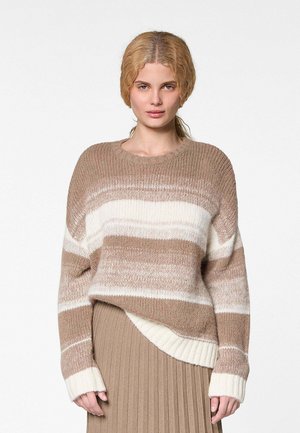 Jonge vrouw met lichtbruin golvend haar draagt een beige en wit gestreepte gebreide trui en een beige geplooide rok tegen een witte achtergrond.