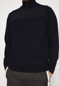 Pull-over noir à manches longues avec demi-fermeture éclair et logo "BOSS" embossé sur la poitrine, porté avec un pantalon foncé avec des bandes blanches sur les côtés.