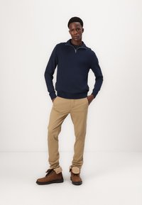 Marineblauer Reißverschluss-Pullover, khakifarbene Hose und braune Stiefeletten mit Futter aus Fell. Das Model steht mit den Händen in den Taschen und zeigt die Details des Outfits.