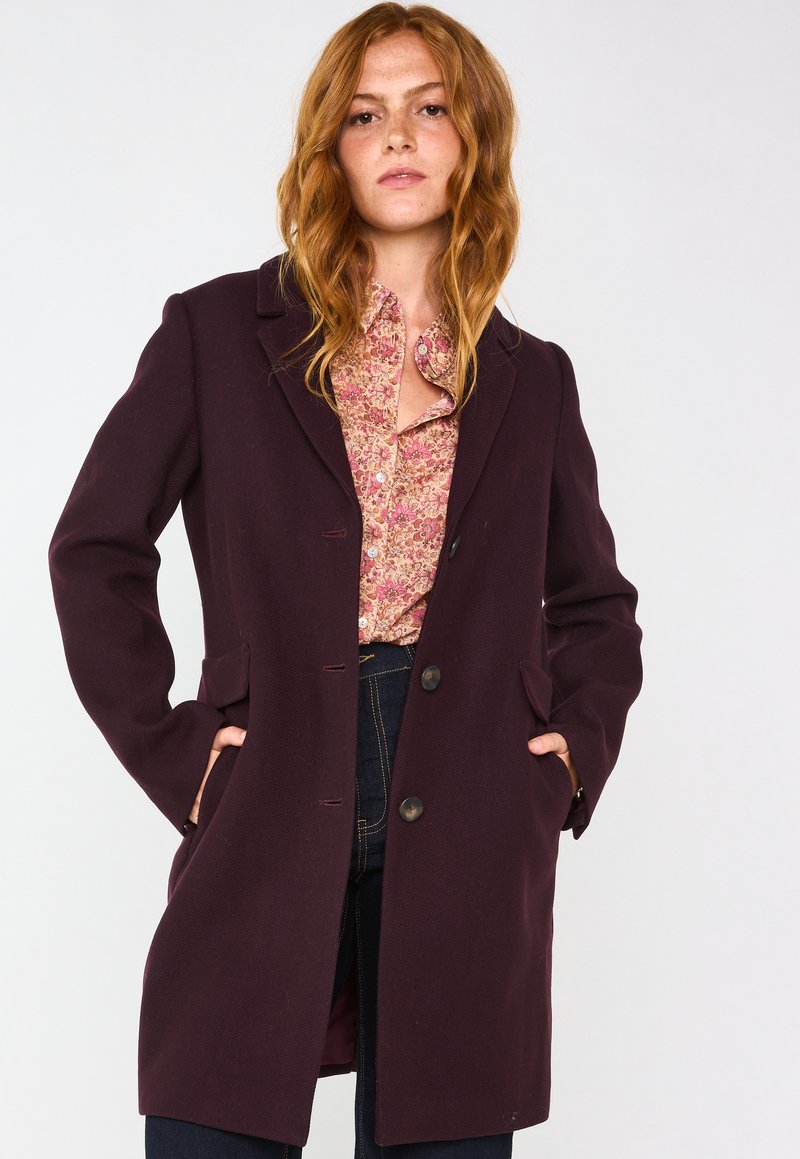 Cappotto di lana viola con un design classico, caratterizzato da un colletto a punta, due tasche frontali e chiusura con bottoni. Tessuto strutturato con una vestibilità sartoriale.