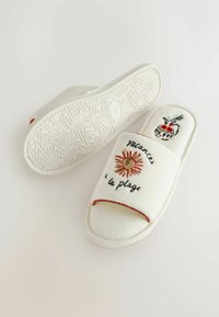 Zapatillas blancas abiertas con bordado de sol, jarrón y texto "vacances à la plage", mostrando suela texturizada y detalle de ribete rojo lateral.