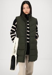 Olivenfarget puffervest med høy krage og sikk-sakk sømmer, brukt over en svart og hvit stripete topp, kombinert med beige bukser.