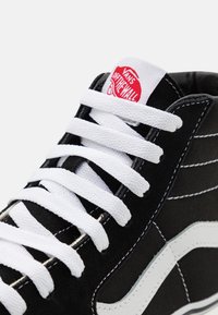 Černé plátěné tenisky s vysokým střihом, bílé šňůrky a švy; mají bílé vlnové logo a červený štítek Vans na jazyku.