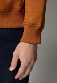 Sudadera naranja con puños acanalados, mostrando una mano descansando sobre un denim oscuro. Enfatiza la textura y el material de la tela y los detalles del ajuste.