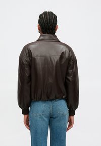 Καφέ δερμάτινο μπουφάν με ψηλό γιακά, φαρδιά εφαρμογή και φουσκωτά μανίκια. Φορεμένο πάνω από ανοιχτόχρωμο τζιν παντελόνι. Ομαλή υφή με λεπτομέρειες ραφής.