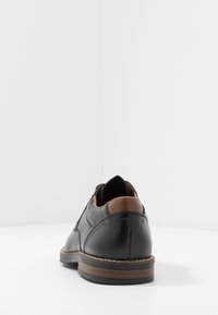 Scarpa in pelle nera con un accento in pelle marrone sul tacco, caratterizzata da dettagli di cucitura e suola in gomma, vista da dietro su sfondo bianco.