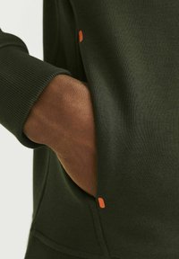Mørkegrønt sweatshirtstof med ribbede manchetter. En hånd hviler i en kengurulomme, der har en orange detalje nær åbningen.