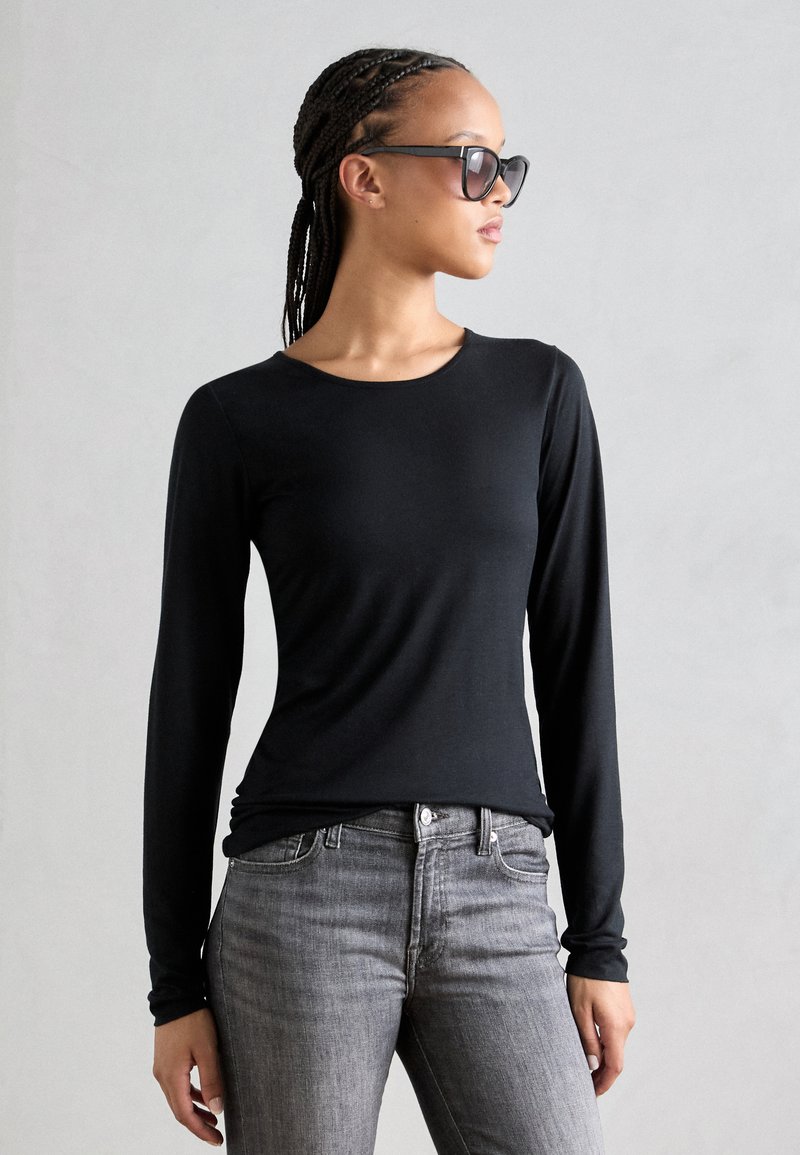 Bluză neagră cu mânecă lungă dintr-un material neted, cu guler rotund și un croi ajustat, purtată împreună cu blugi gri din denim.