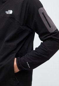 Μανίκι μαύρου μπουφάν The North Face με τσέπη με φερμουάρ στο πάνω μέρος του βραχίονα και κείμενο "FUTUREFLEECE" κοντά στον καρπό, χέρι στην πλαϊνή τσέπη.