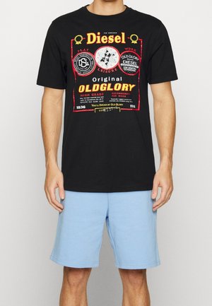 Mies, joka seisoo vaaleansinisissä shortseissa ja mustassa Diesel Oldglory -graafisessa t-paidassa yksinkertaisen taustan edessä.