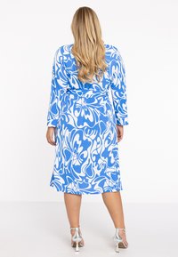 Blaues und weißes Kleid mit Muster, langen Ärmeln, einem gebundenen Taillenteil und einem fließenden Rock. Strukturierter Stoff bringt visuelles Interesse. Silberne Absätze.