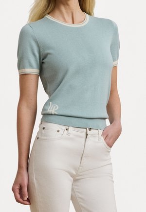 Femme portant un haut tricoté bleu clair à manches courtes avec bord blanc et un jean blanc taille haute, main dans la poche, sur fond uni.