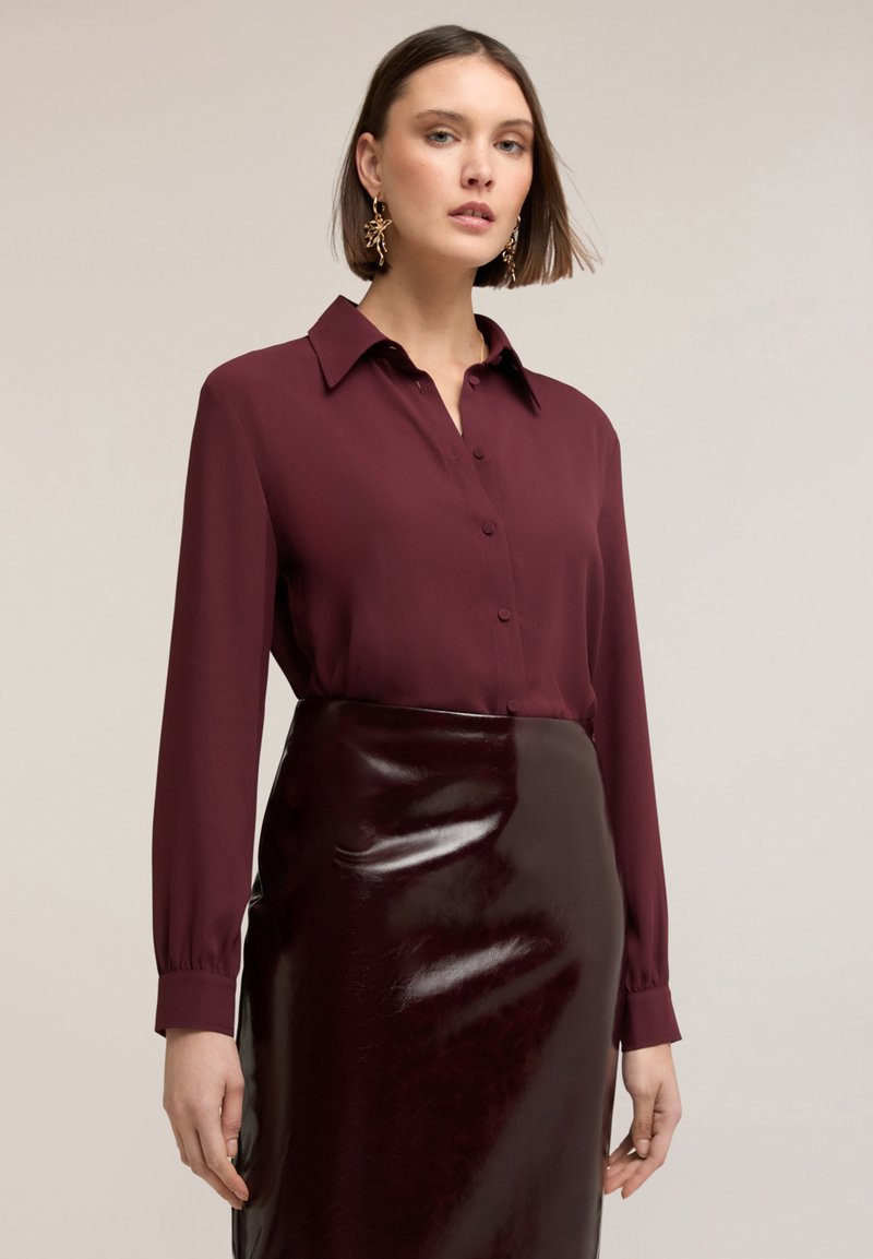 Kobieta w bordowej bluzce zapinanej na guziki i błyszczącej ciemnoczerwonej spódnicy z winylu na tle jasnego jednolitego tła.