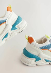 Białe sneakersy z niebieskimi, pomarańczowymi i neonowymi zielonymi akcentami. Posiadają teksturowaną podeszwę i siateczkowy wierzch z wyraźnymi kształtami i wzorami.