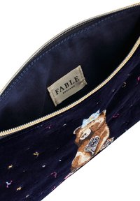 Blå sammetspåse med en guldfärgad dragkedja, med en broderad björndesign. Insidan av etiketten visar "FABLE" i svart text.