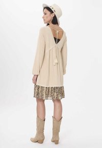 Beige jurk met lange mouwen en een lage rug, voorzien van kantaccenten en een rok met slangenhuidprint. Gecombineerd met beige cowboystiefels.