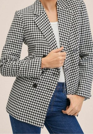 Vrouw in een zwart-witte ruitblazer over een wit shirt en blauwe jeans, die met één hand de blazer recht trekt.