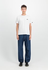 Balts kokvilnas T-krekls ar mazu melnu logotipu kreisajā krūšu daļā, apvienots ar brīvu melnā zila denim biksēm un melnām kurpēm.