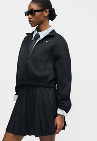 Γυναίκα με μαύρο μπουφάν με φερμουάρ, πλισέ φούστα, γαλάζιο πουκάμισο, μαύρη γραβάτα και μαύρα γυαλιά ηλίου σε απλό φόντο.