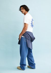 Witte t-shirt met blauw grafisch ontwerp, gecombineerd met losse denim jeans en een blauwgestreepte blouse geknoopt om de taille. Beige instap schoenen.