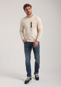 Sudadera beige con puños y cuello acanalados, que presenta un bolsillo en el pecho con cremallera negra. Combinada con vaqueros azules y zapatillas negras.
