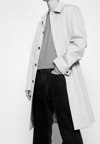 Cappotto beige chiaro realizzato in tessuto morbido, con colletto, bottoni neri e una vestibilità ampia. Indossato sopra un maglione grigio e pantaloni in corduroy scuri.