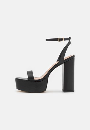 Zwarte hoge platform sandalen met een brede strap aan de voorkant, enkelbandje en een stevige blokhak. Luxe leren textuur, minimalistisch ontwerp.