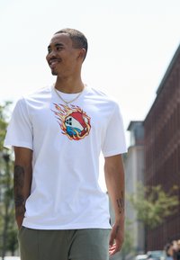 T-shirt alb cu mânecă scurtă, având un grafic colorat cu o flacără și un design stilizat. Purtați-l cu pantaloni verzi deschis, în aer liber, cu clădiri în fundal.